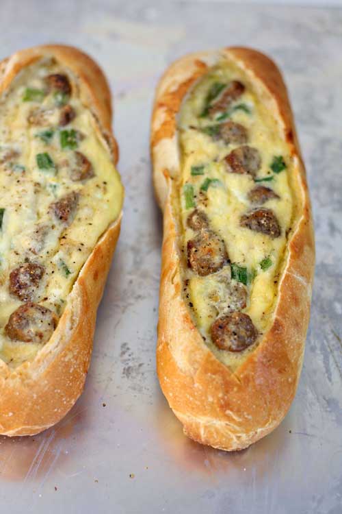 Two sourdough baguettes that have been split open along the top. Each is filled with sausage, green onion, and baked eggs.