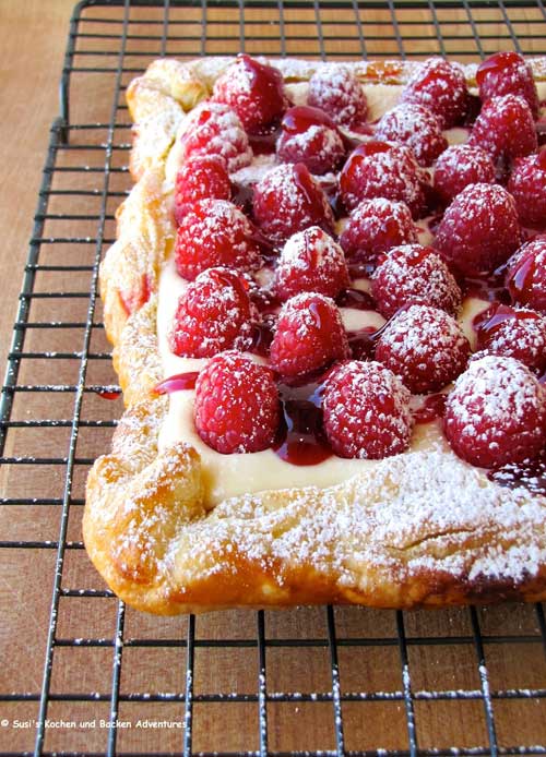 A rectangular tart covered in raspberries rests on a wire rack. The top of the tart has been sprinkled with powdered sugar.