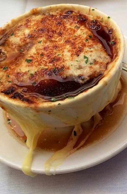 A white cup filled with French onion soup. The soup is covered with melted cheese, that is running down the side of the cup and onto a white plate that the cup is resting on.