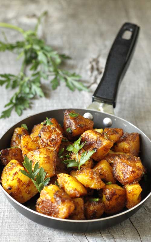 Roasted Bombay Potatoes in a small skillet with a black handle. The skillet is sitting on grey, weathered wood. Fresh cilantro  leaves garnish the potatoes, and a full spring sits in the background.