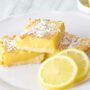 Prepared lemon bars and lemon slices on a white plate. The bars have been dusted with powdered sugar.